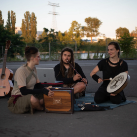 Drei Musizierende sitzen auf einem Teppich. Zu sehen sind verschiedene Instrumente wie z.B. Shrutibox, Kaval, Rahmentrommel und Gitarre.