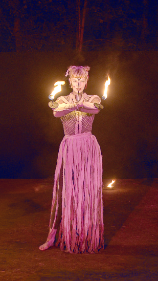 Clara Komann als Klotho, einer der drei Moiren, singend mit zwei Handflammen.